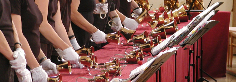 Mehrere Handglocken liegen auf einem langen Tisch, dahinter die Hände und Arme der Musikantinnen