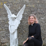 Pfarrerin Theilacker-Dürr kniet in schwarzer Amztskleidung neben einer weißen Engels-Statue.