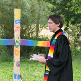 Pfarrer Simon Dürr im Talar steht im Freien vor einem bunten Holzkreuz.